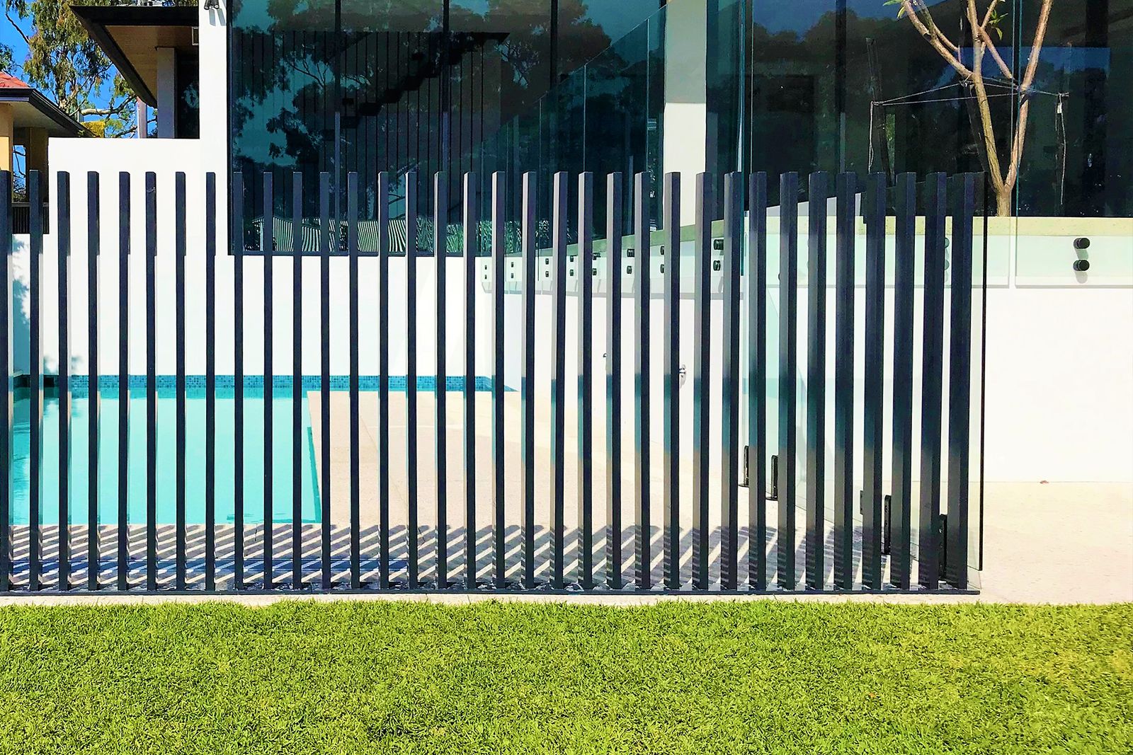 A charcoal aluminium pool barrier on lawn, with a frameless glass fence and turquoise water behind