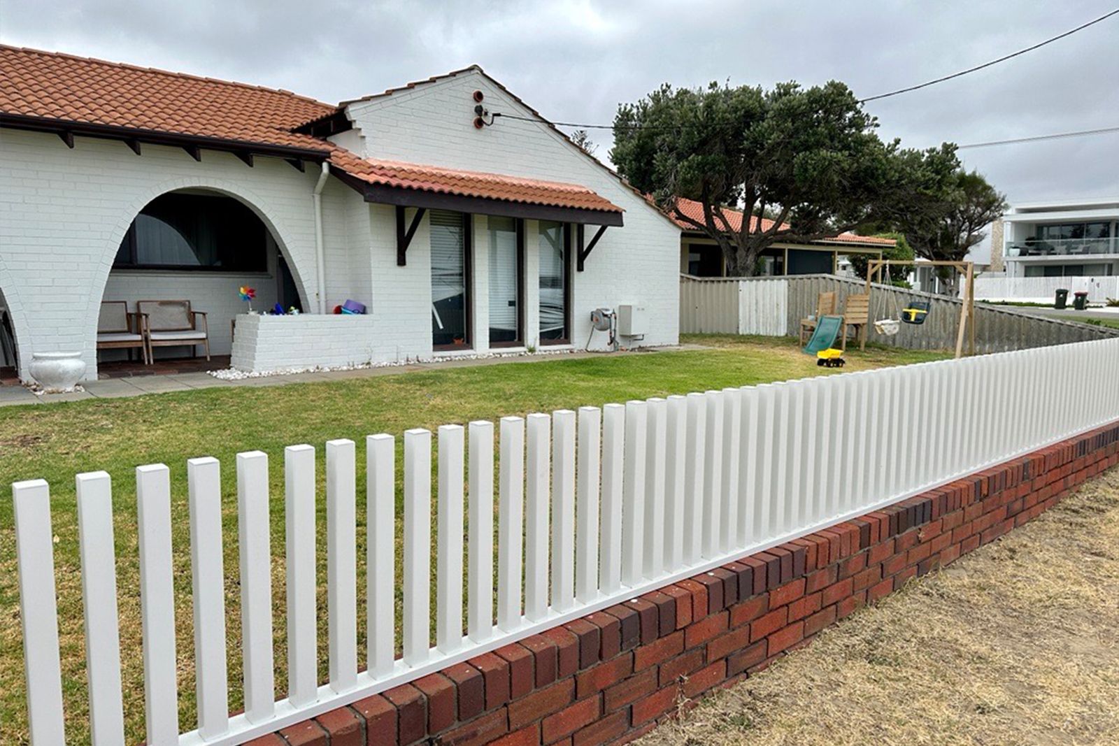 A white aluminium fence on a red brick plinth in front of a white brick house with a terracotta roof