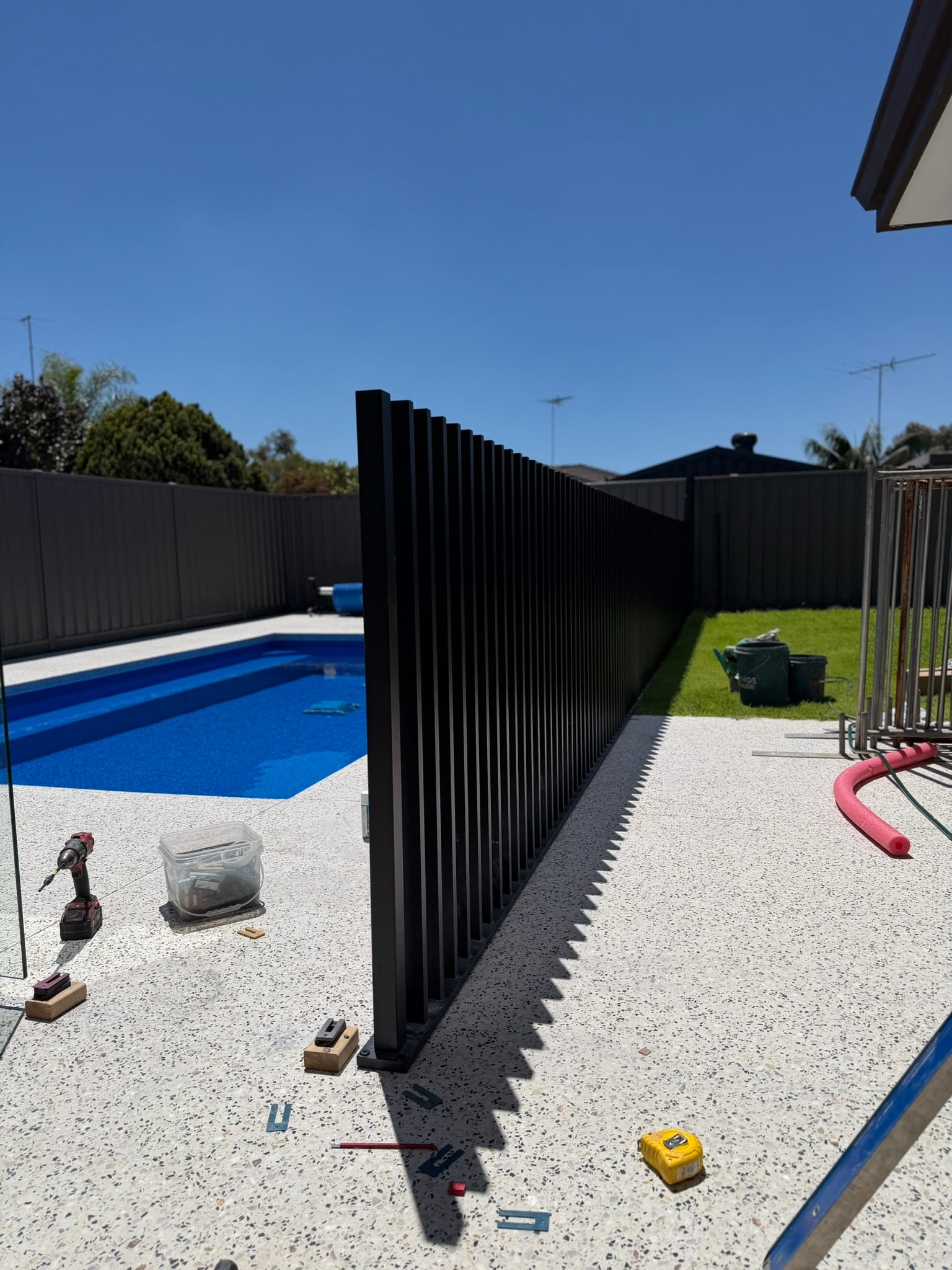 A fence run part way through installation, with a drill, fixings and a tape measure on the paving